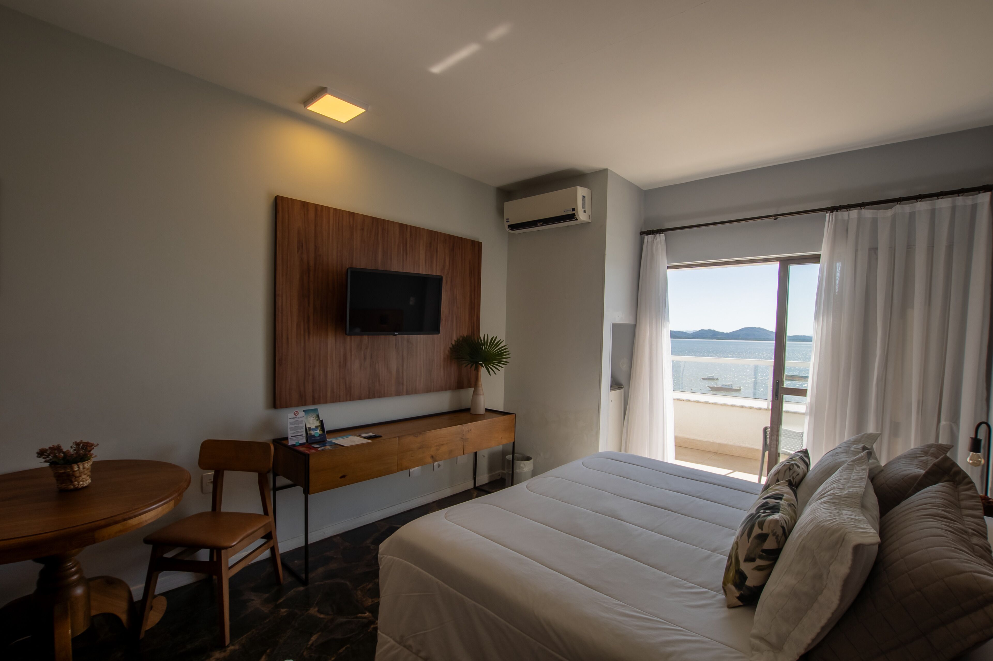 Een hotelkamer met een bed, een bureau, een aan de muur bevestigde televisie, een kamerplant en uitzicht op zee.