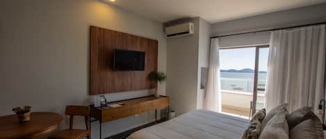 A hotel room with a bed, a desk, a TV mounted on the wall, a potted plant, and a view of the sea.
