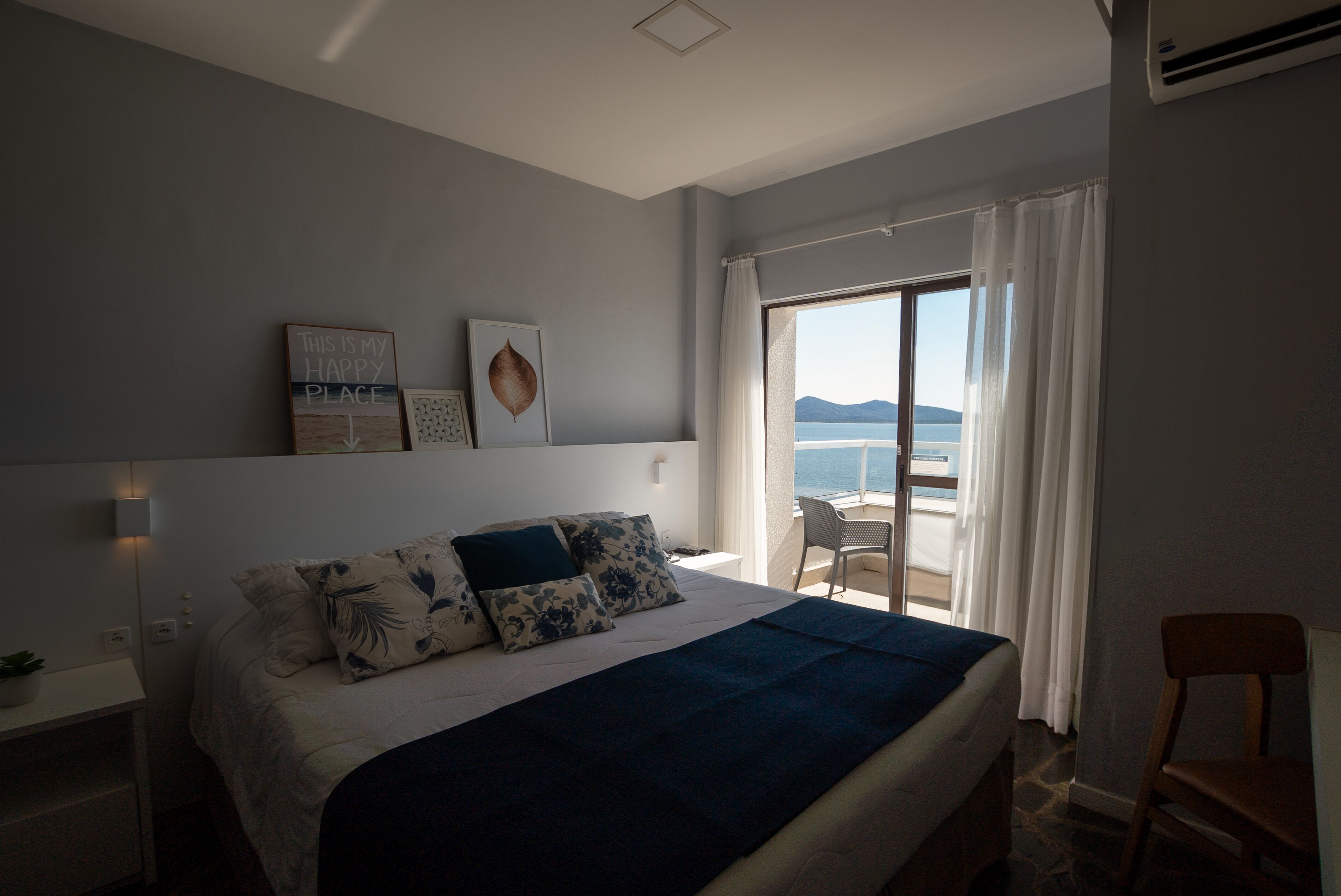 A bedroom with a bed, a nightstand, a chair, and a view of the sea.