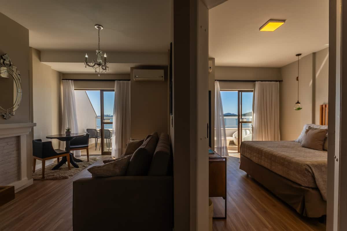 A modern hotel room with a bed, a sofa, a dining area with a table and chairs, a chandelier, and a view of the sea.