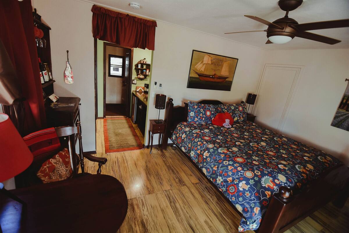 A bedroom with a wooden floor, a bed with a floral bedspread, a ceiling fan, and a door leading to another room.