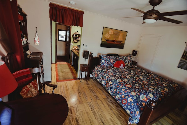Un dormitorio con piso de madera, una cama con cubrecama floral, un ventilador de techo y una puerta que da a otra habitación.