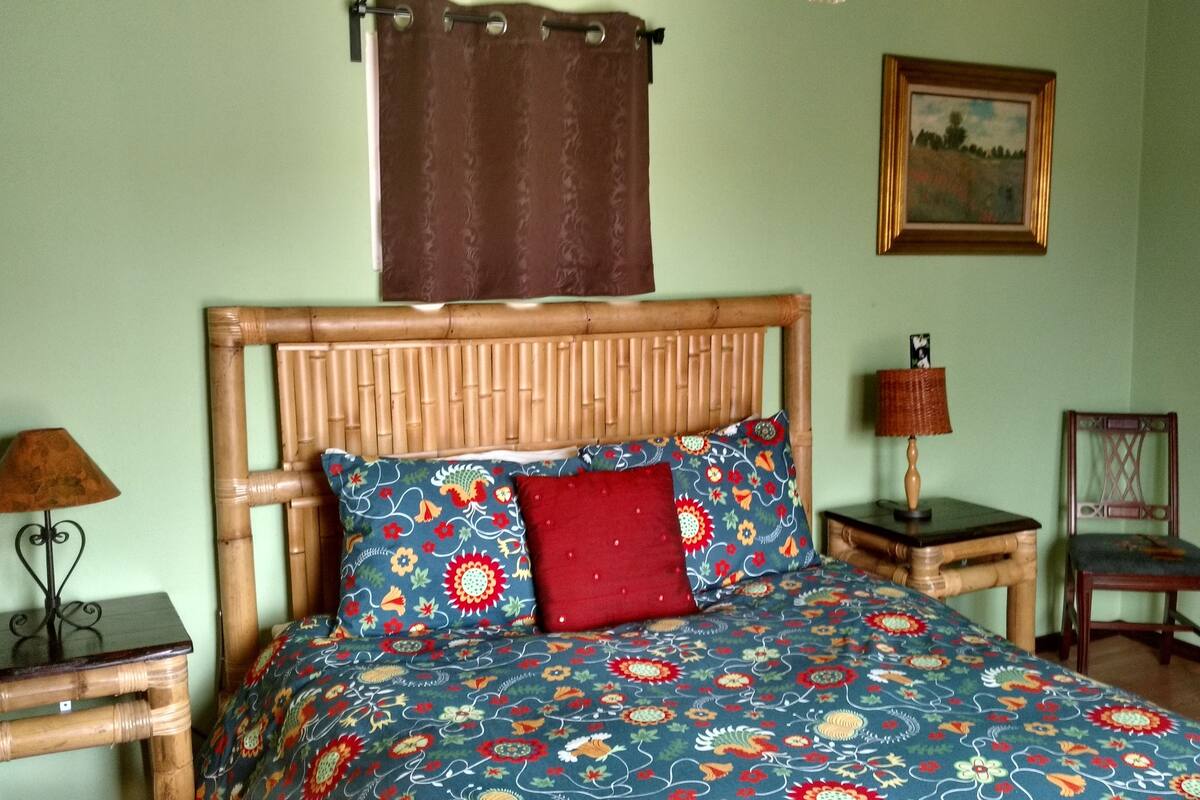 A bed with a patterned bedspread, a wooden headboard, a red pillow, a nightstand with a lamp, a chair, and a framed picture on the wall.