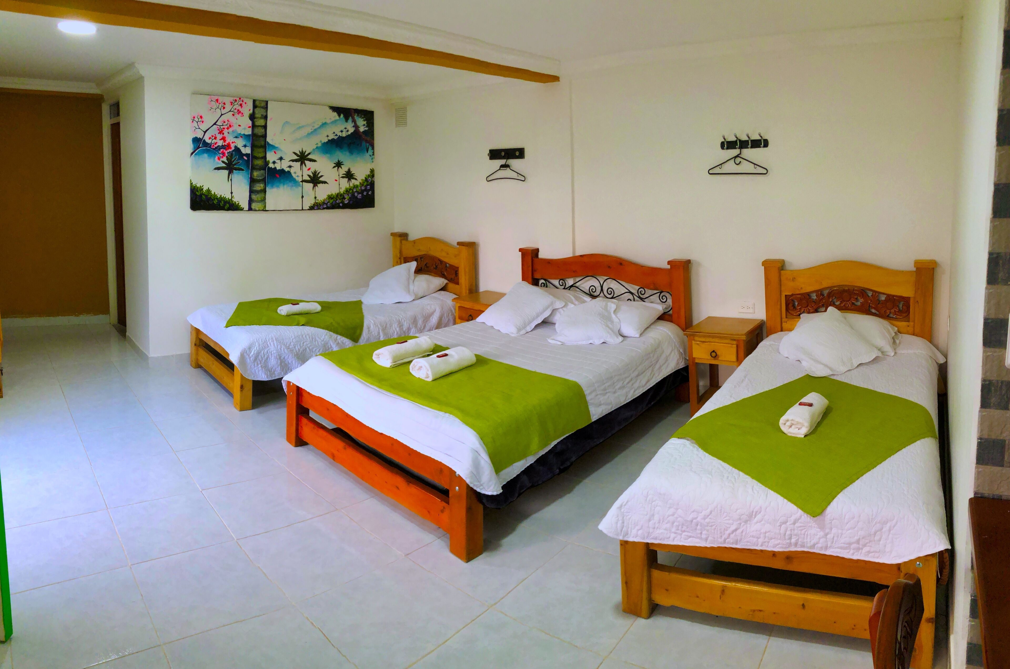 A hotel room with three wooden beds, each with a green and white bedspread and white pillows. There are two wall-mounted hooks and a painting on the wall.
