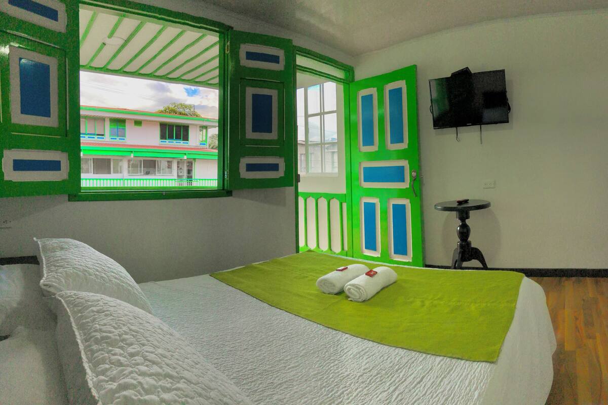 Ein Zimmer mit einem Bett, einem grünen Fenster, einem Fernseher an der Wand und einem kleinen Tisch.
