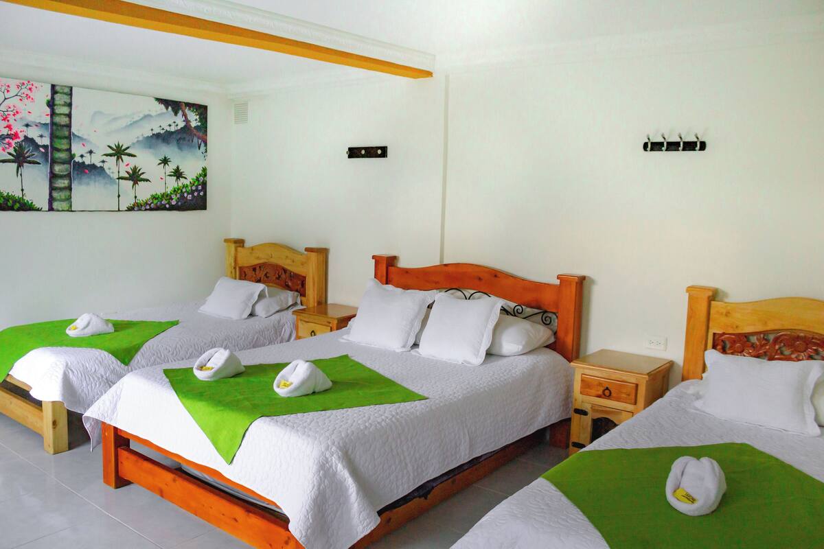 Ein Hotelzimmer mit drei Holzbetten, jedes mit weißer Bettwäsche und grünen Handtüchern. An der Wand hängt ein Gemälde und ein kleiner Nachttisch aus Holz mit einer Schublade.
