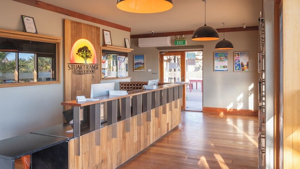 A reception desk with a wooden finish, a sign for Stuart Range Outback Resort, and a view of the outdoor area through the window. - Stuart Range Outback Resort (Coober Pedy)