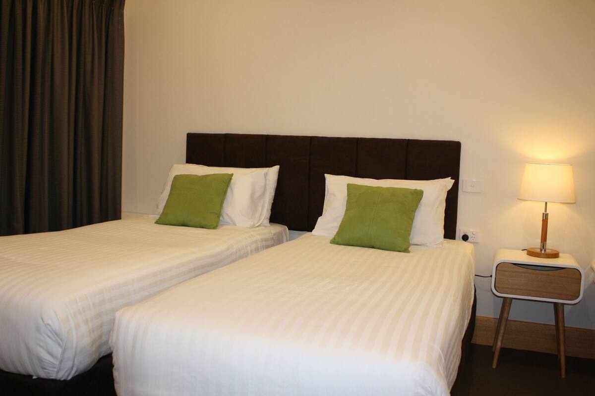 A hotel room with two single beds, each with white bedding and green pillows. There is a bedside table with a lamp and a power outlet.