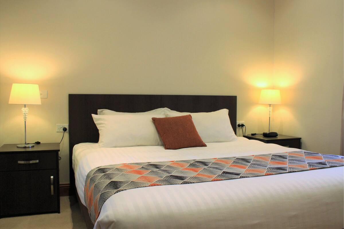 A neatly made bed with a patterned bedspread, two bedside lamps, and a dark headboard.