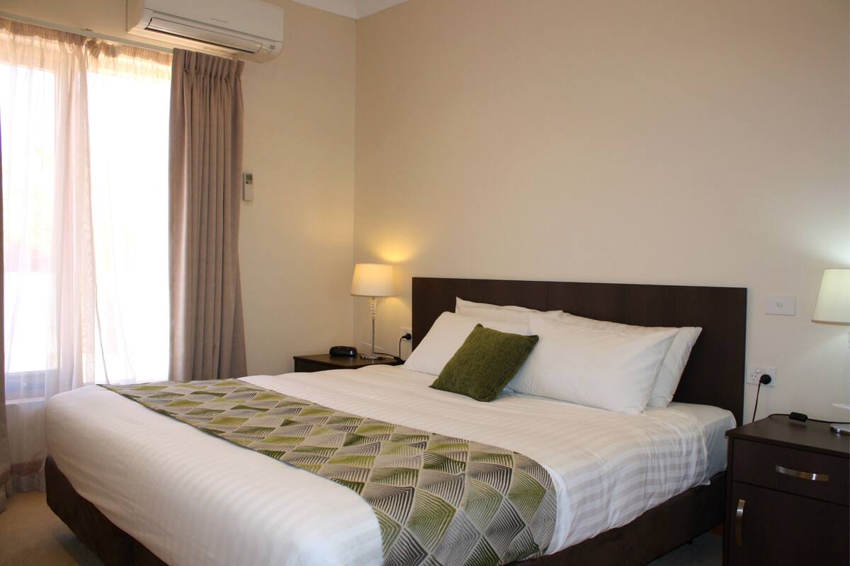 A neatly made bed with a patterned bedspread, a bedside table with a lamp, and a window with curtains.