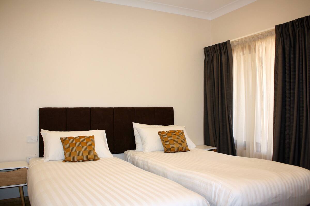 A hotel room with two beds, a dark headboard, white bedding, and a window with dark curtains.