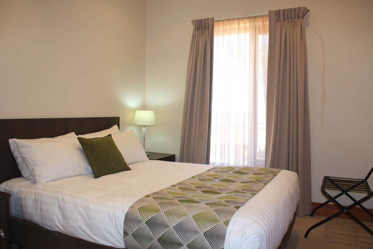A neatly made bed with a patterned bedspread, a bedside lamp, and a chair in a room with a large window and curtains.