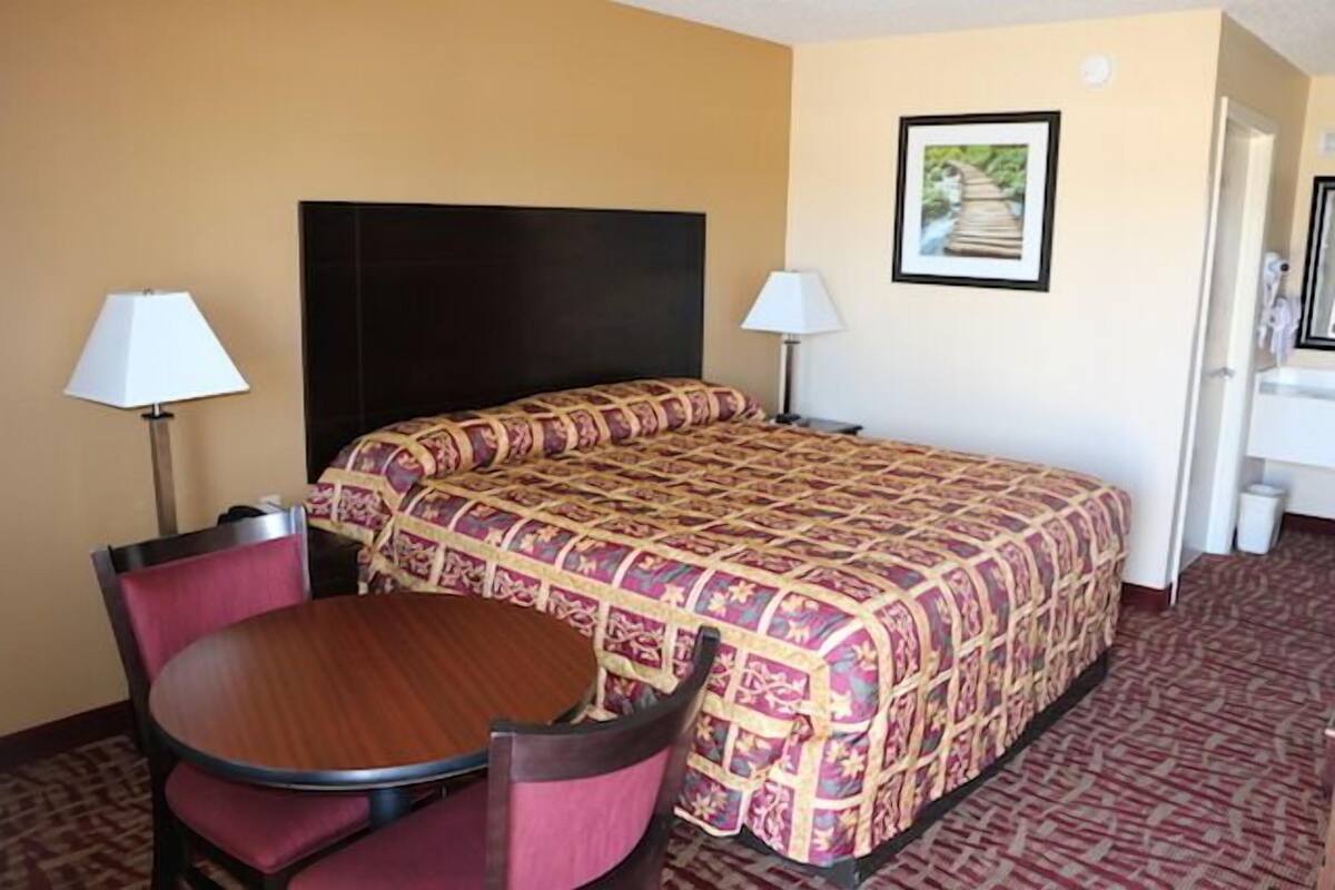 A hotel room with a bed, two bedside lamps, a round table with chairs, and a framed picture on the wall.