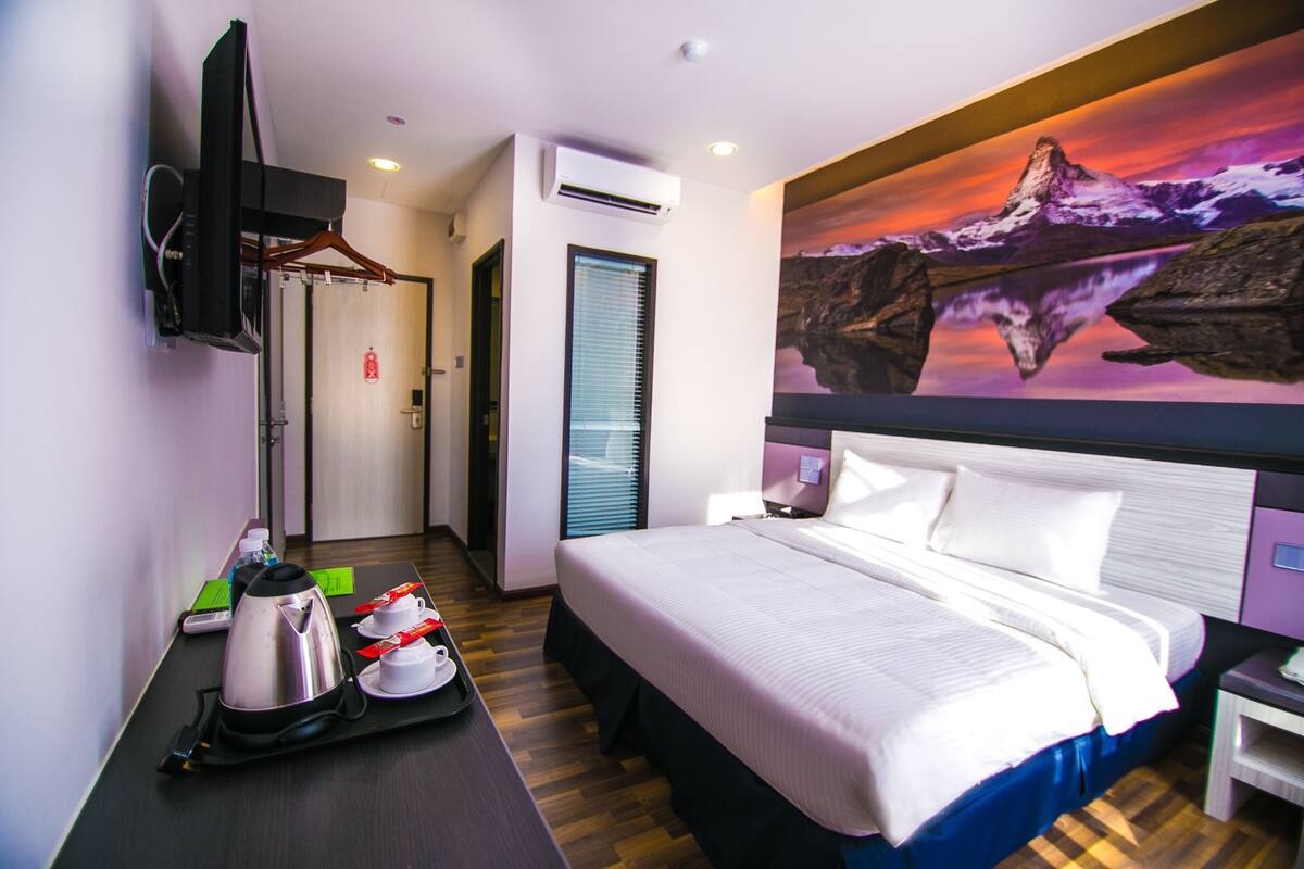 Une chambre d’hôtel avec un grand lit, un téléviseur à écran plat fixé au mur, un appareil de climatisation, un tableau représentant un paysage de montagne et un bureau avec un plateau contenant des articles pour le petit-déjeuner.