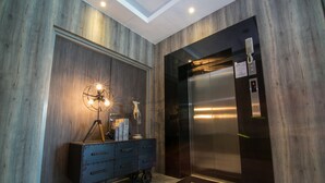 Un ascensor moderno con pantalla digital y panel de control, una pared de madera con una lámpara decorativa, y un carrito de equipaje azul con felpudo.