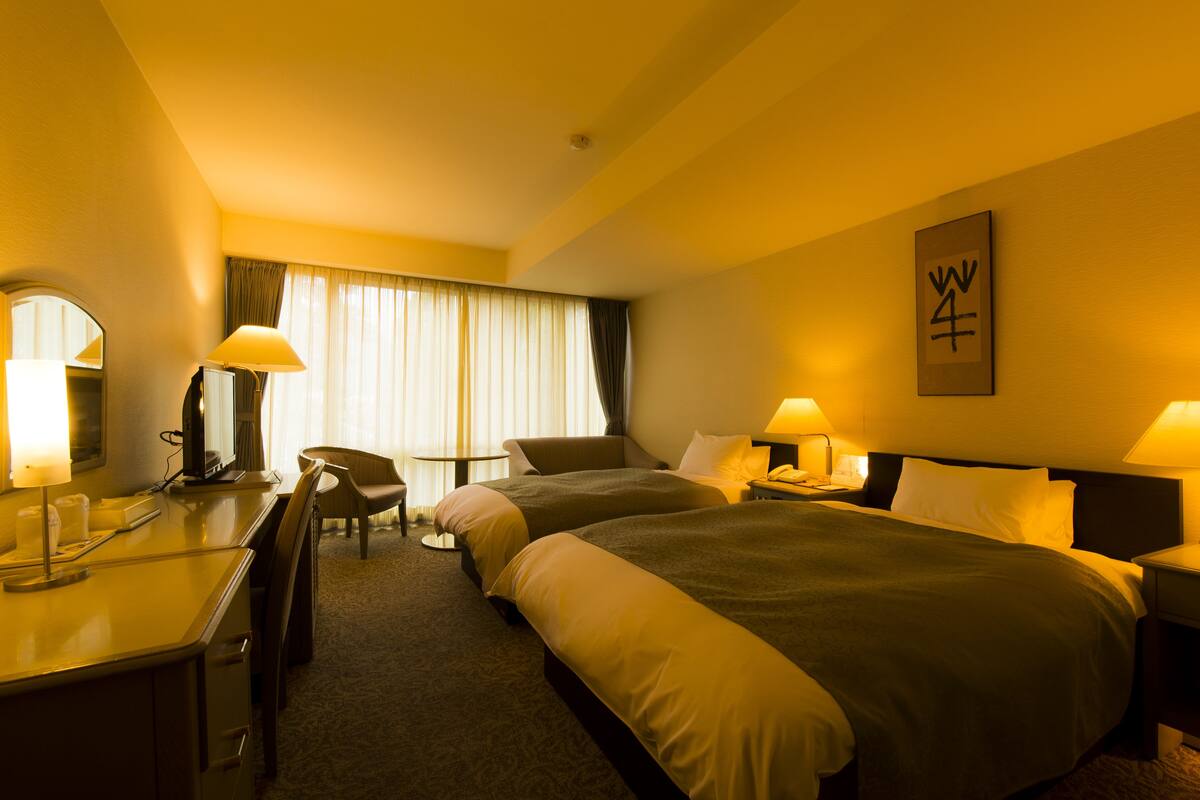 A hotel room with two beds, a desk, a chair, a desk lamp, a bedside table with a lamp, a large window with curtains, and a wall-mounted sign.