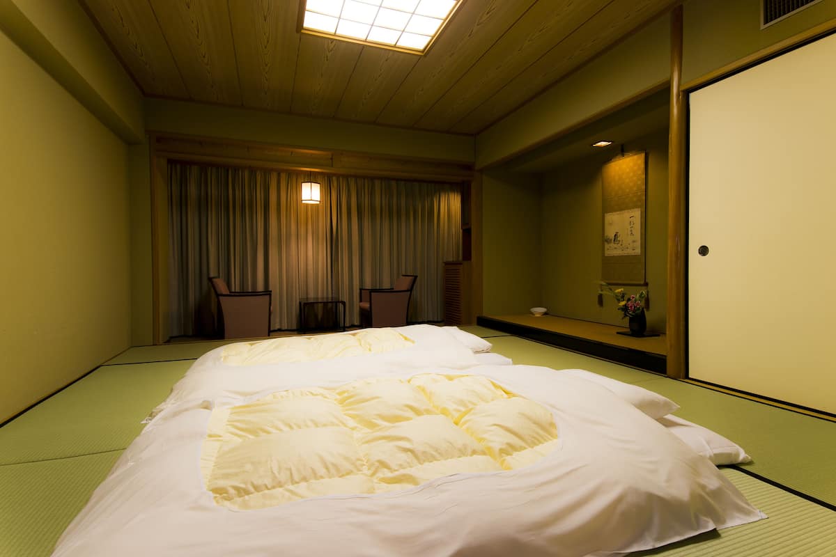 A traditional Japanese room with tatami flooring, a low bed with white bedding, a sliding door, and a small table with chairs.