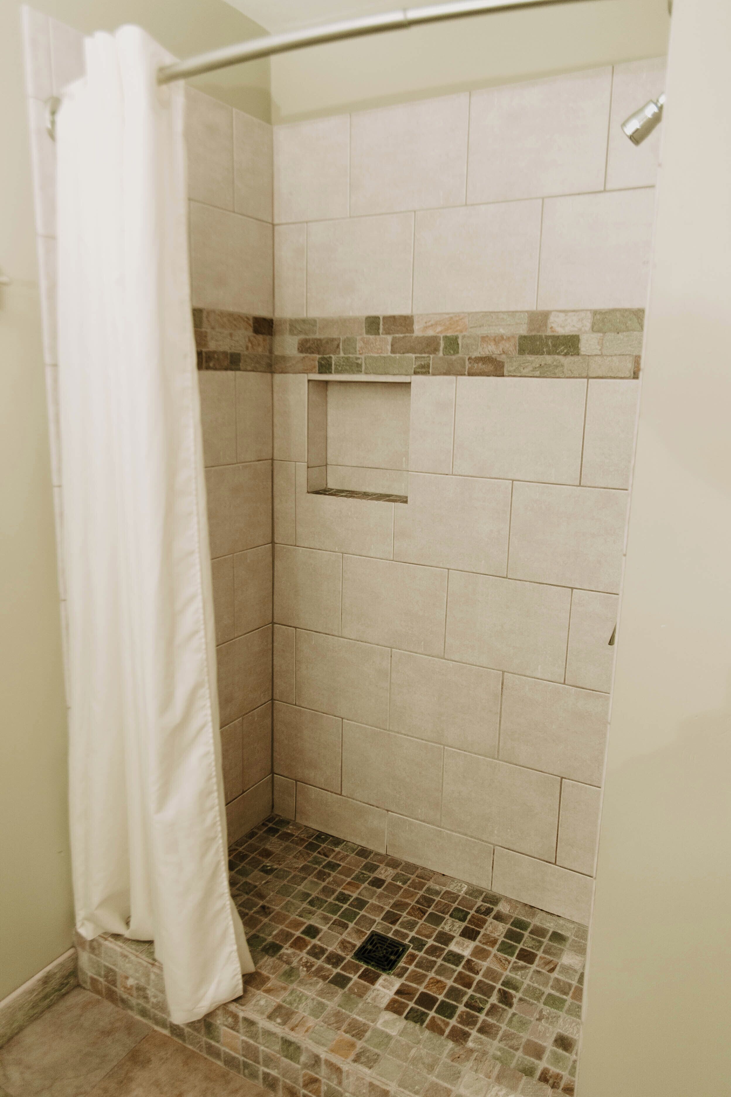 A shower area with beige tiles, a white shower curtain, and a small shelf.