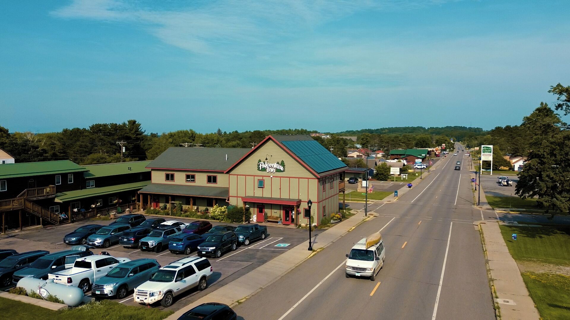 A two-story building with a green roof and a sign reading "Adventure Inn." There is a parking lot with various cars and a van on the street.