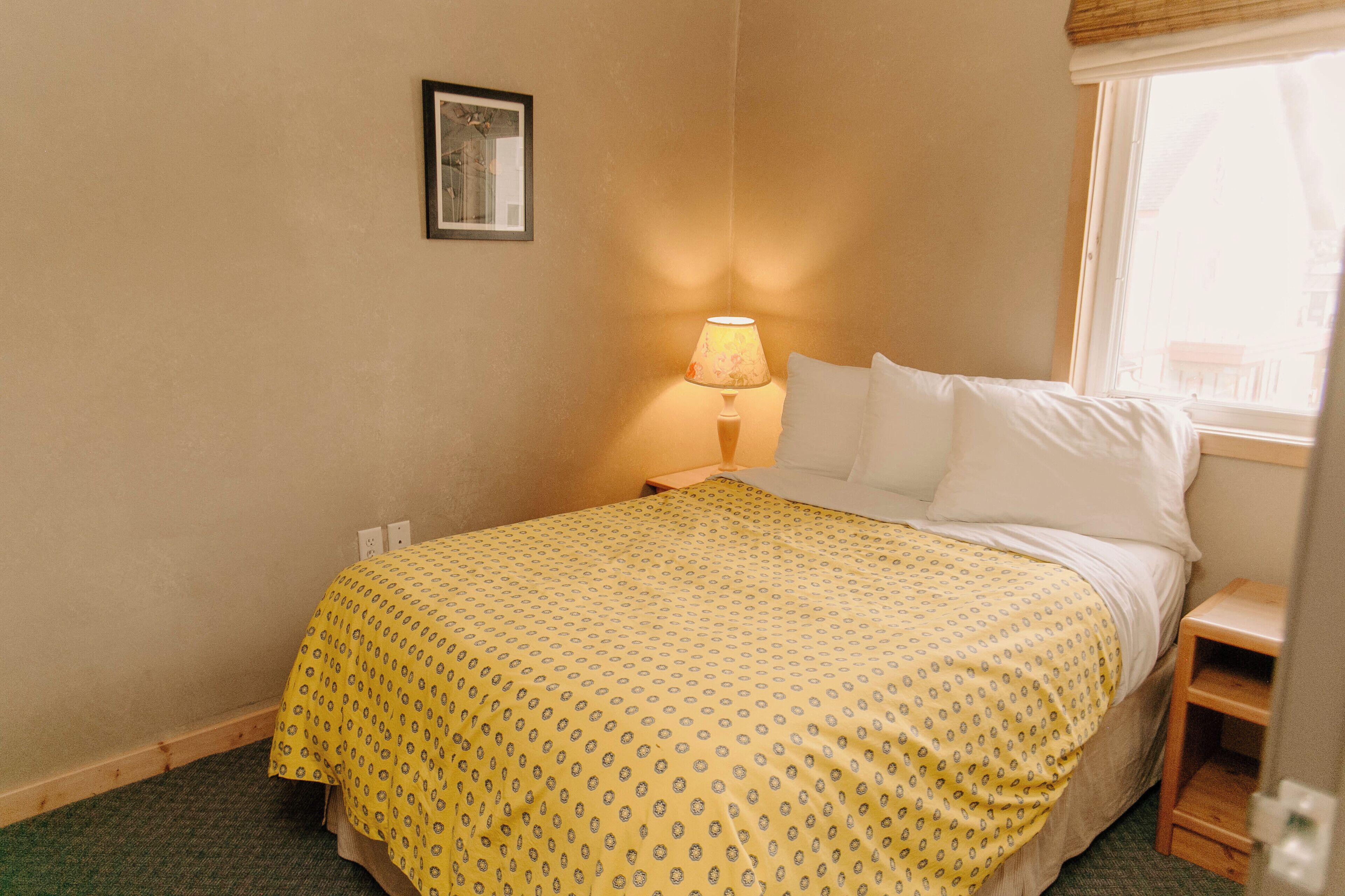 Una habitación de hotel con una cama, una mesita de noche, una lámpara, una ventana y un cuadro en la pared.