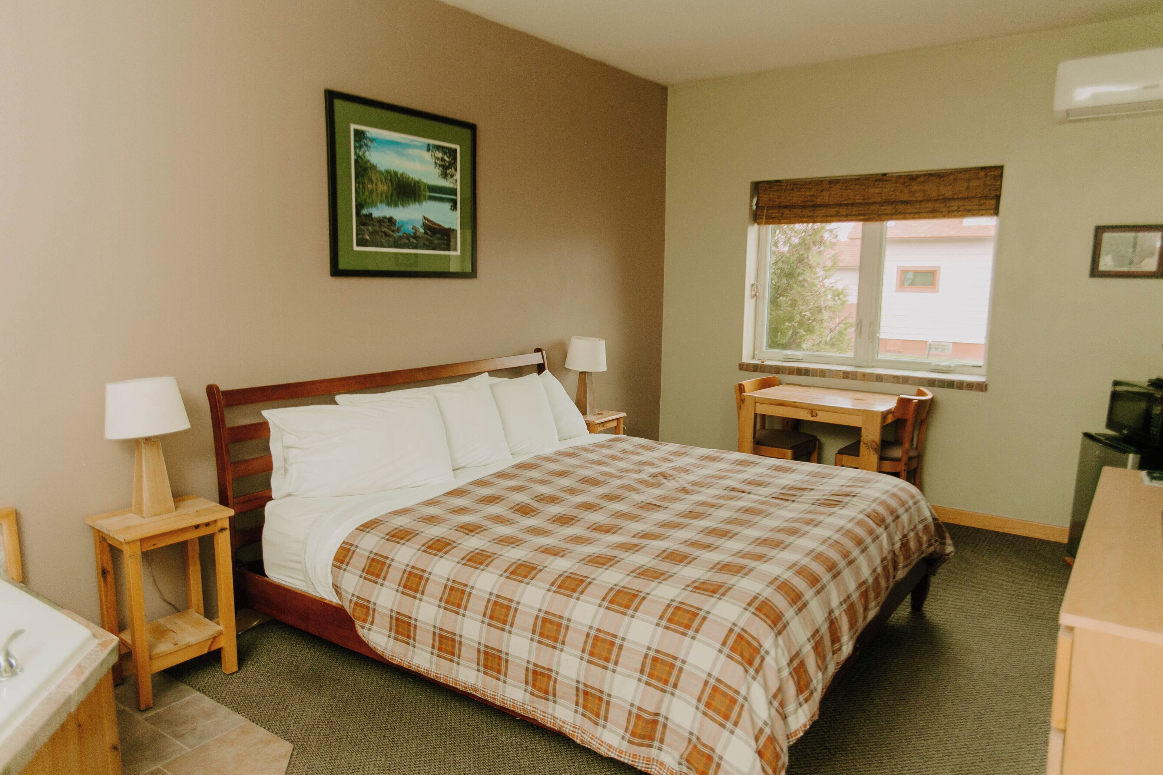 Une chambre d’hôtel avec un lit, un bureau, une chaise, une lampe, un tableau au mur et une fenêtre.