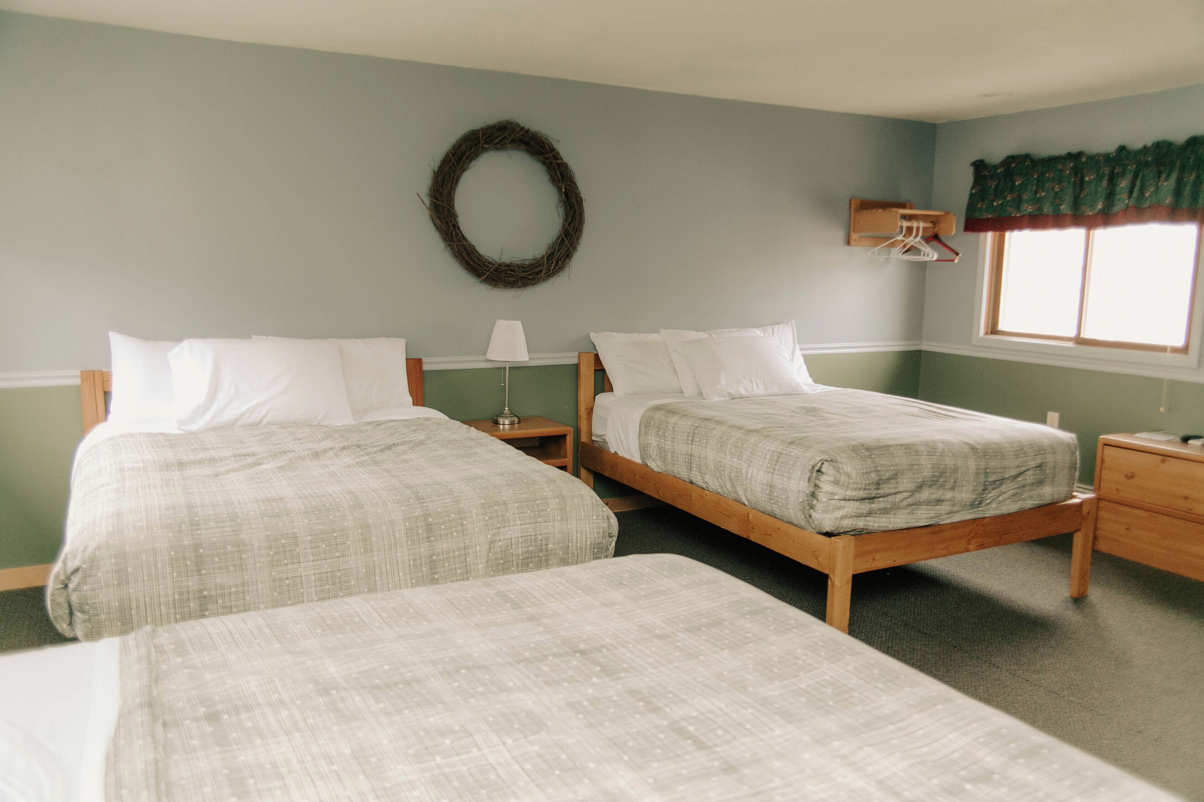 A room with two single beds, a wooden headboard, a small table with a lamp, a wall-mounted coat rack, and a window with a green valance.
