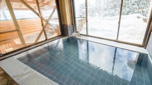 An indoor swimming pool with clear glass walls, a wooden deck outside, and a snowy landscape visible through the windows. - Bergtour Marukita (Hakuba)