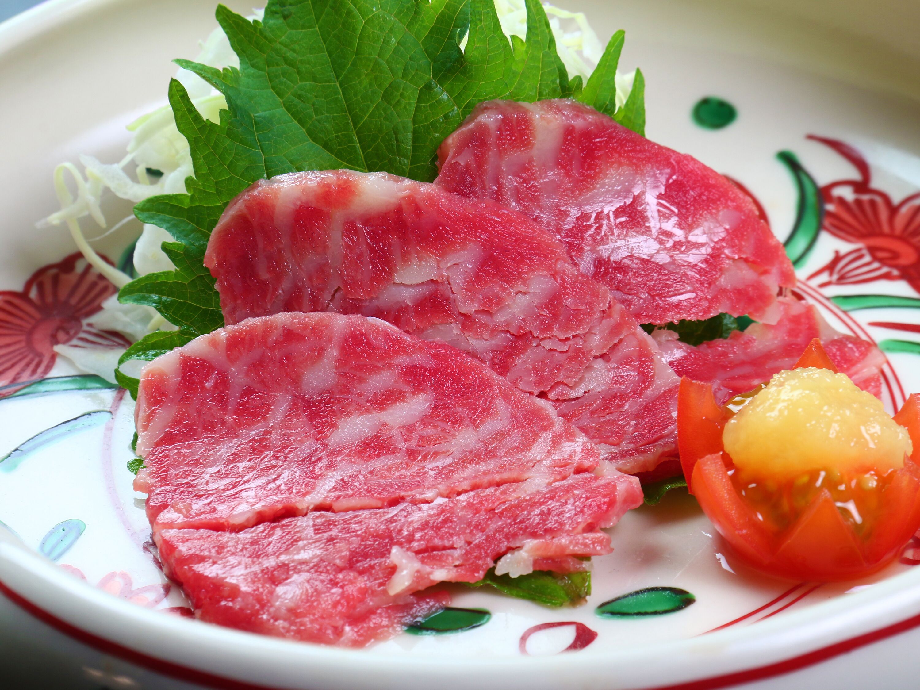 A plate with thinly sliced meat, a small tomato, and green leaves.