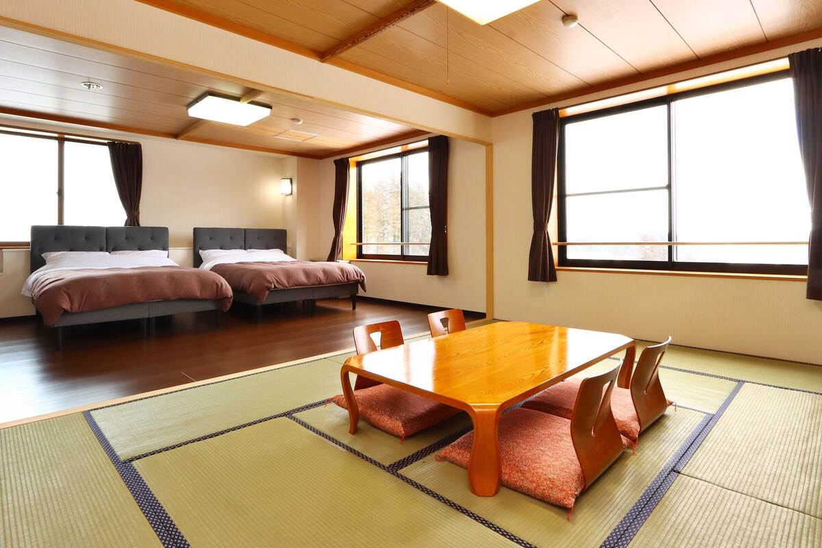 A hotel room with two beds, a dining table, tatami mats, and large windows.