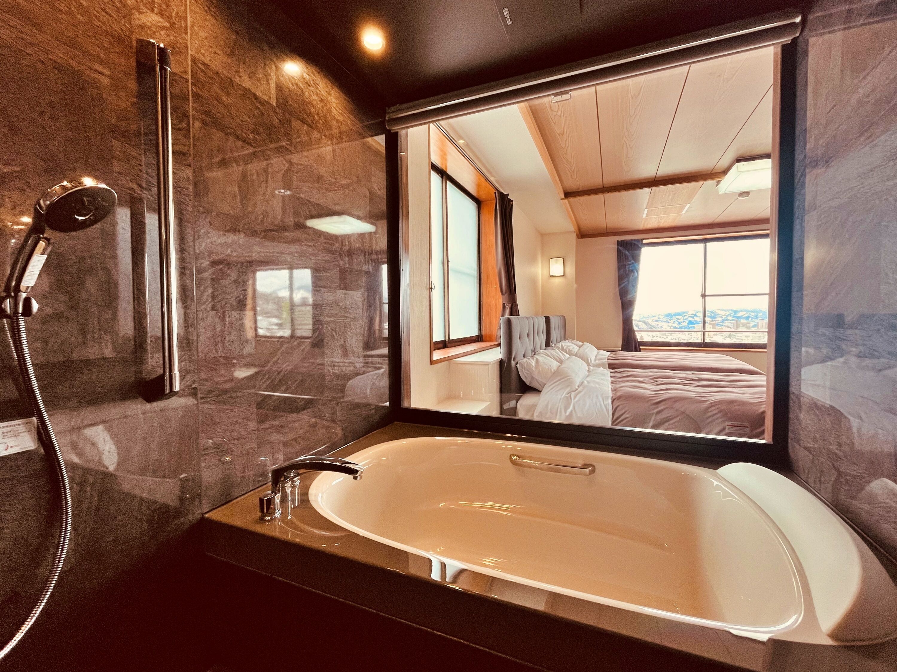 Une salle de bain moderne dotée d’une grande baignoire, d’une douche et d’un miroir qui reflète une chambre à coucher avec un lit et une fenêtre.