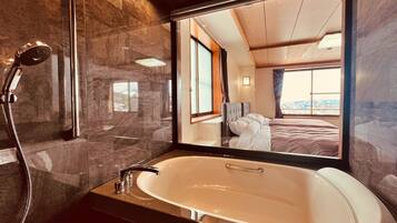 Une salle de bain moderne dotée d’une grande baignoire, d’une douche et d’un miroir qui reflète une chambre à coucher avec un lit et une fenêtre.