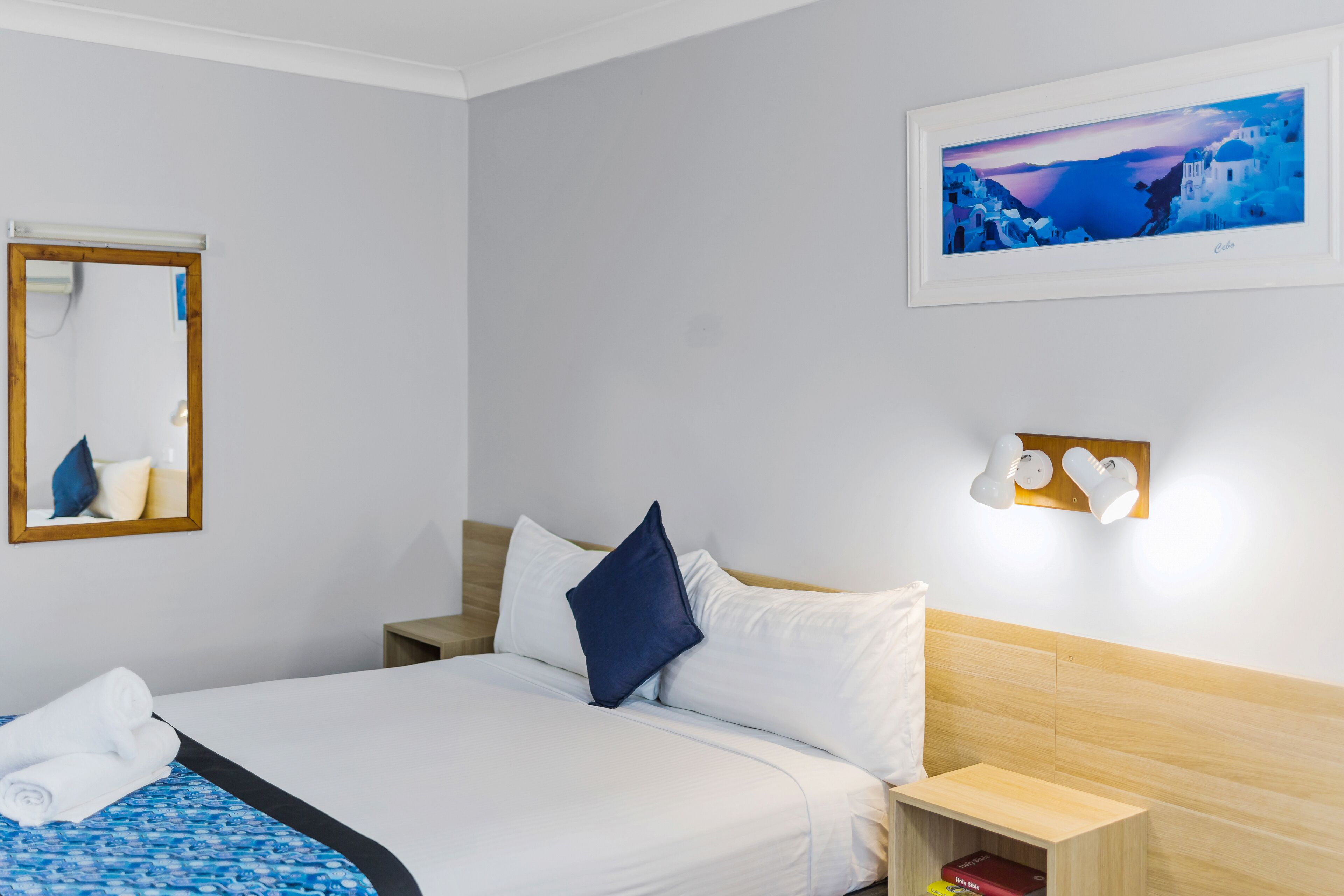 A hotel room with a bed, a nightstand, a bedside table, a mirror, and a framed picture on the wall.