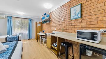A compact room with a kitchenette, a bed with blue bedding, a wooden desk, and a brick wall.