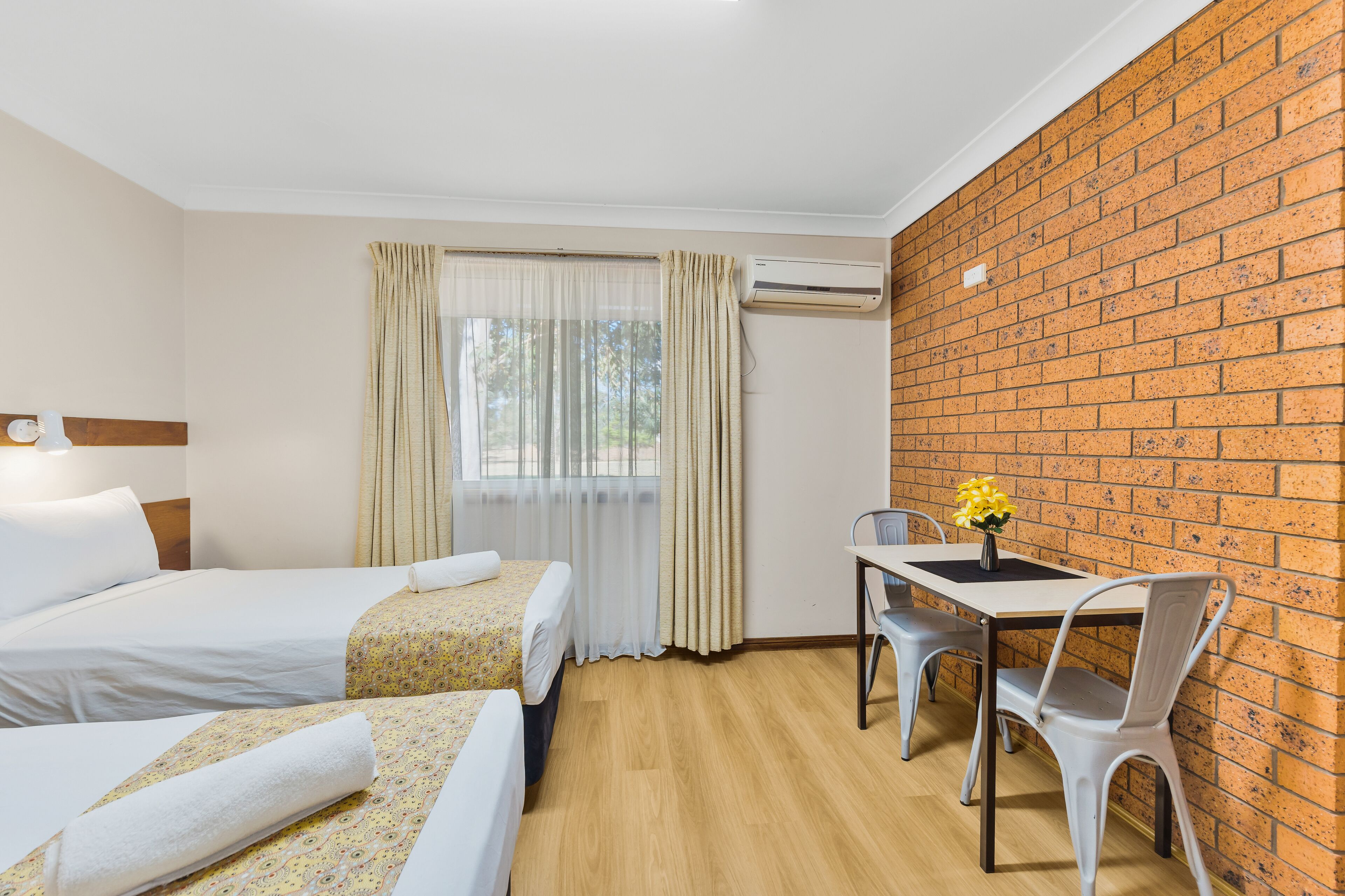 Habitación de hotel con dos camas, una mesa de comedor con sillas, un equipo de aire acondicionado y una pared de ladrillo.