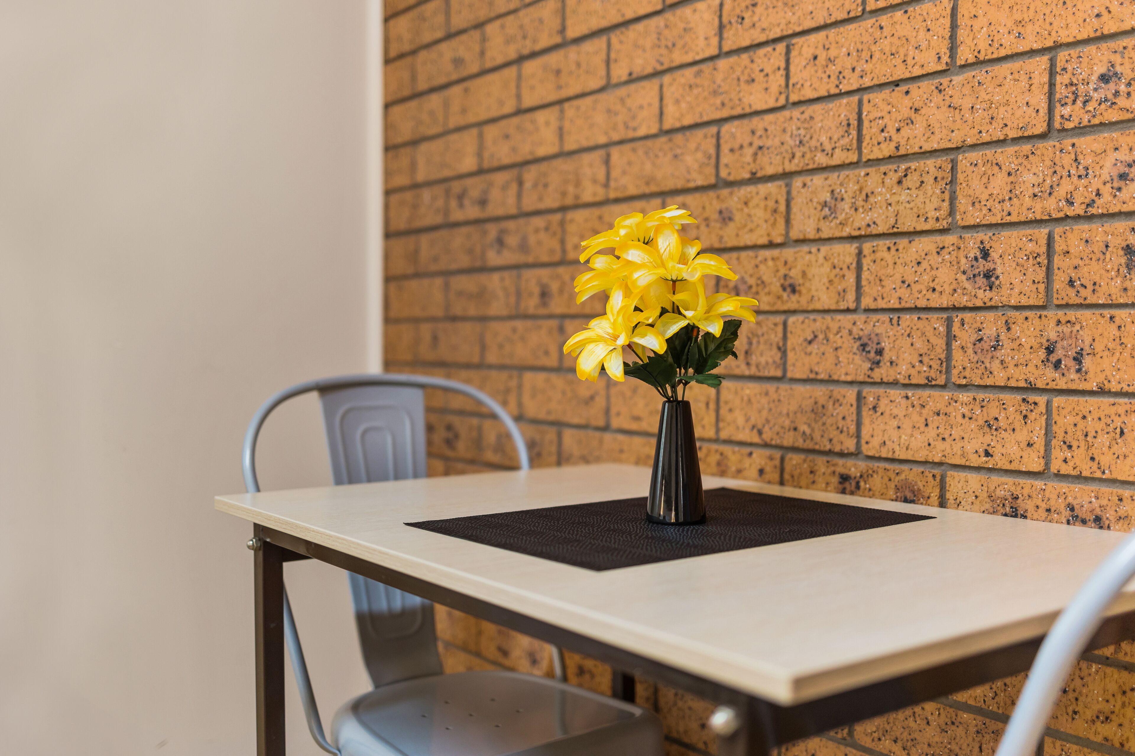 Een tafel met een vaas gele bloemen, een stoel en een bakstenen muur op de achtergrond.