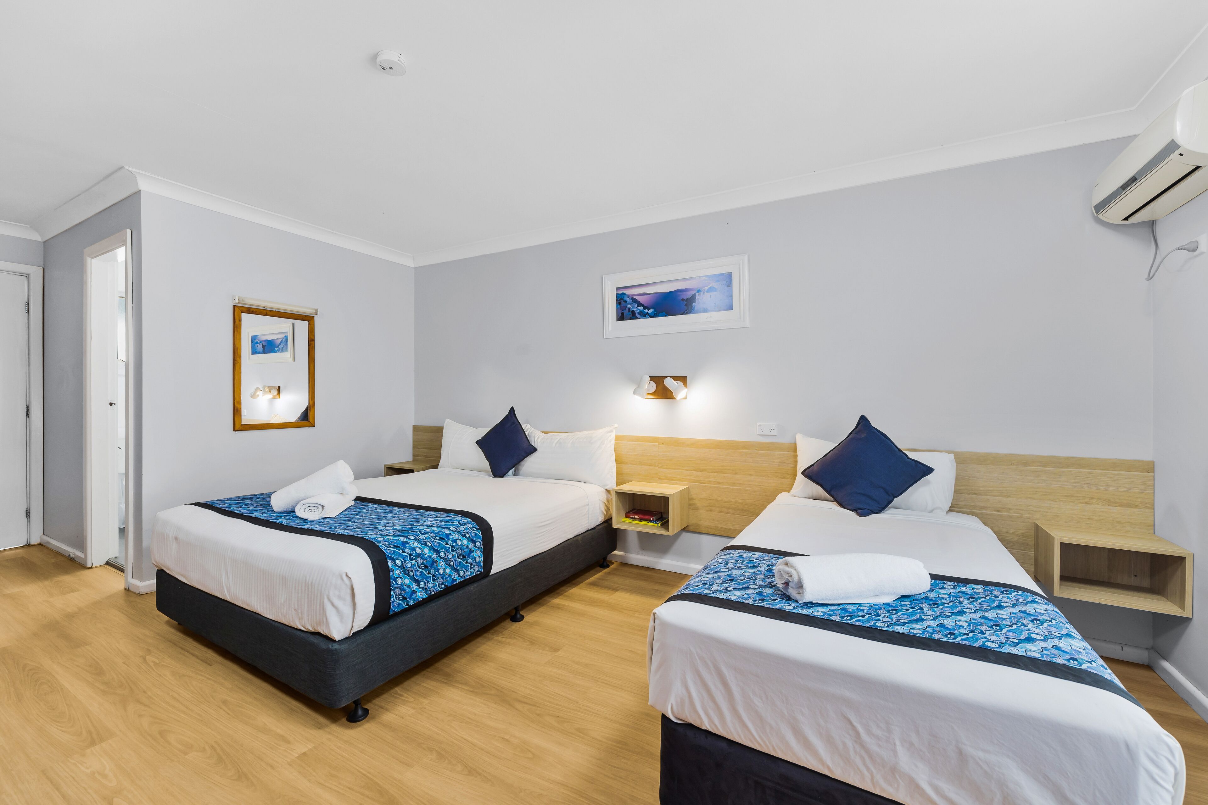 A hotel room with two beds, a wooden headboard, a nightstand, a bedside table, and a wall-mounted air conditioner.