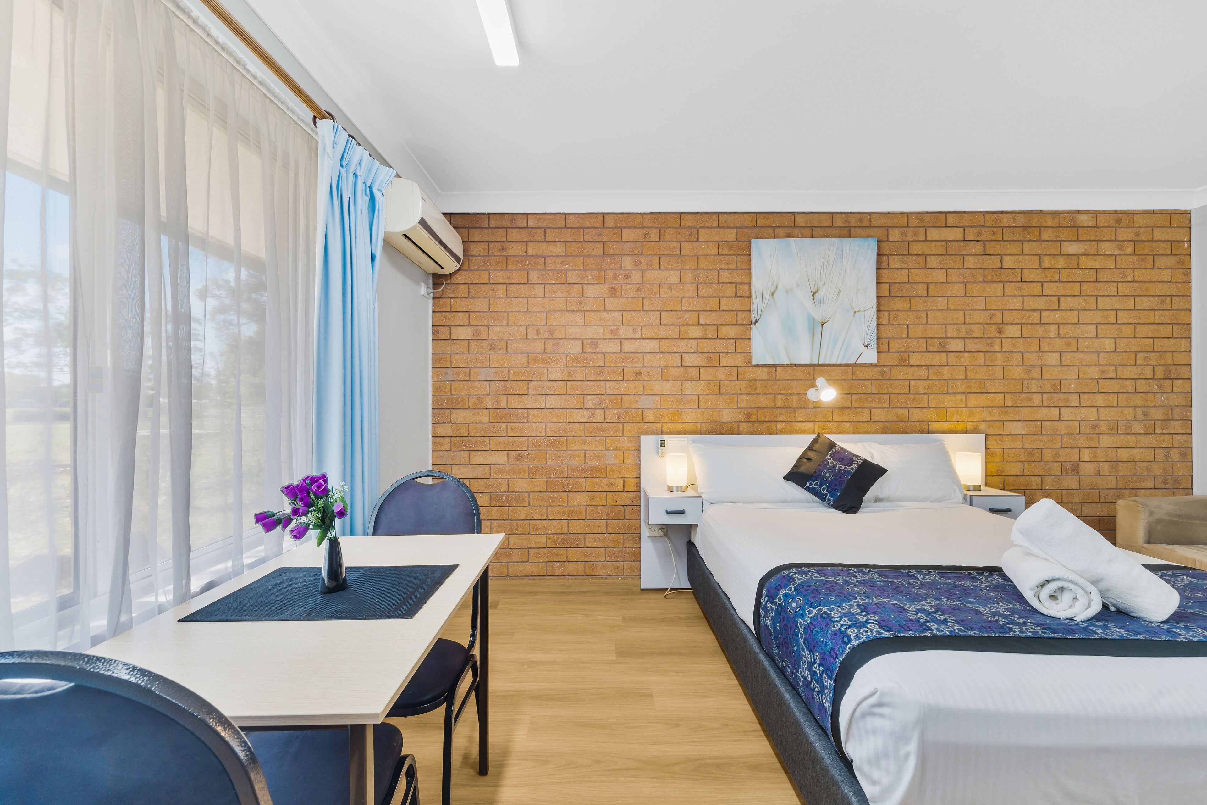 Habitación de hotel con pared de ladrillo, cama con colcha azul estampada, escritorio con silla y ventana con cortinas traslúcidas.