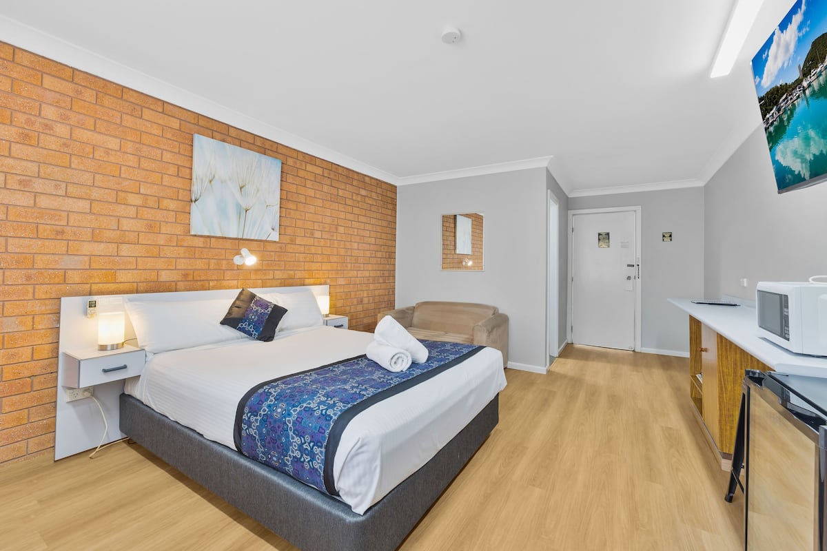 A hotel room with a brick accent wall, a bed with a patterned bedspread, a bedside table with a lamp, a chair, a desk with a microwave, and a door leading to another room.