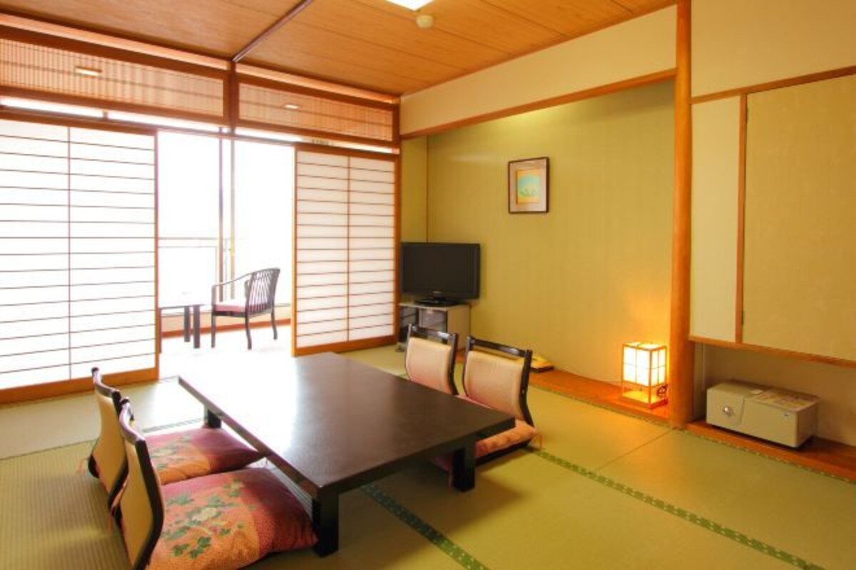 A traditional Japanese room with tatami flooring, a low table, chairs, a television, and a small bedside table with a lamp.