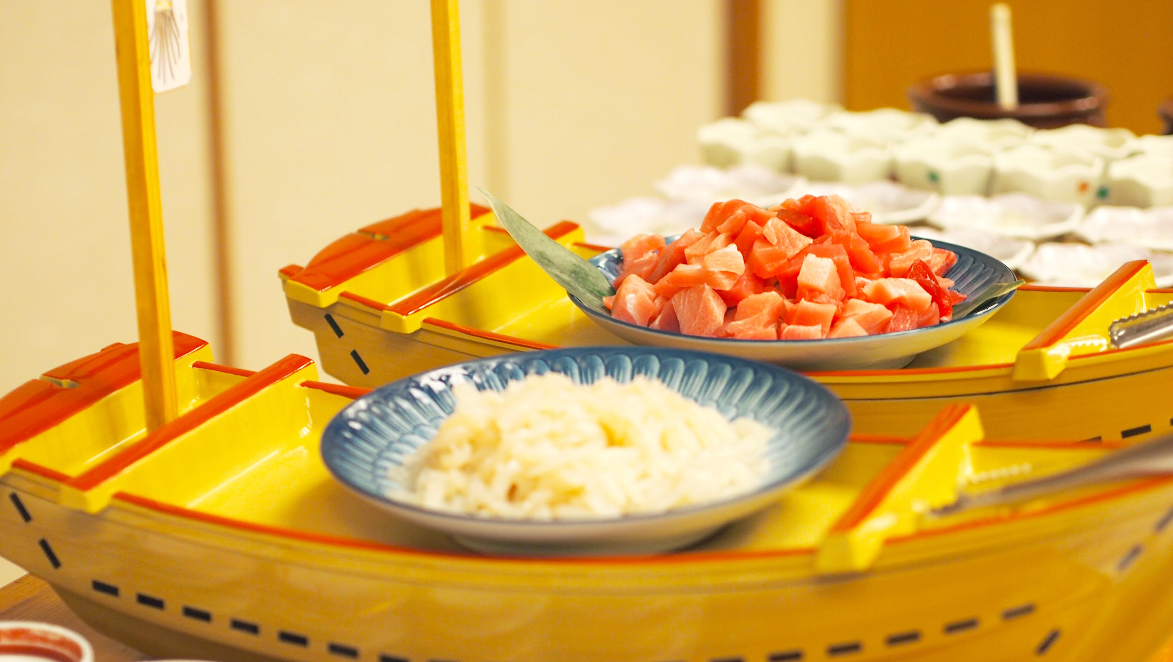 A table with two yellow boats containing bowls of food, one with diced carrots and the other with diced watermelon.
