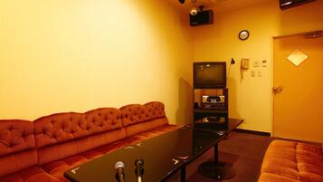 Une chambre d’hôtel avec un canapé capitonné, une petite table sur laquelle se trouve un microphone, une télévision et une horloge murale.