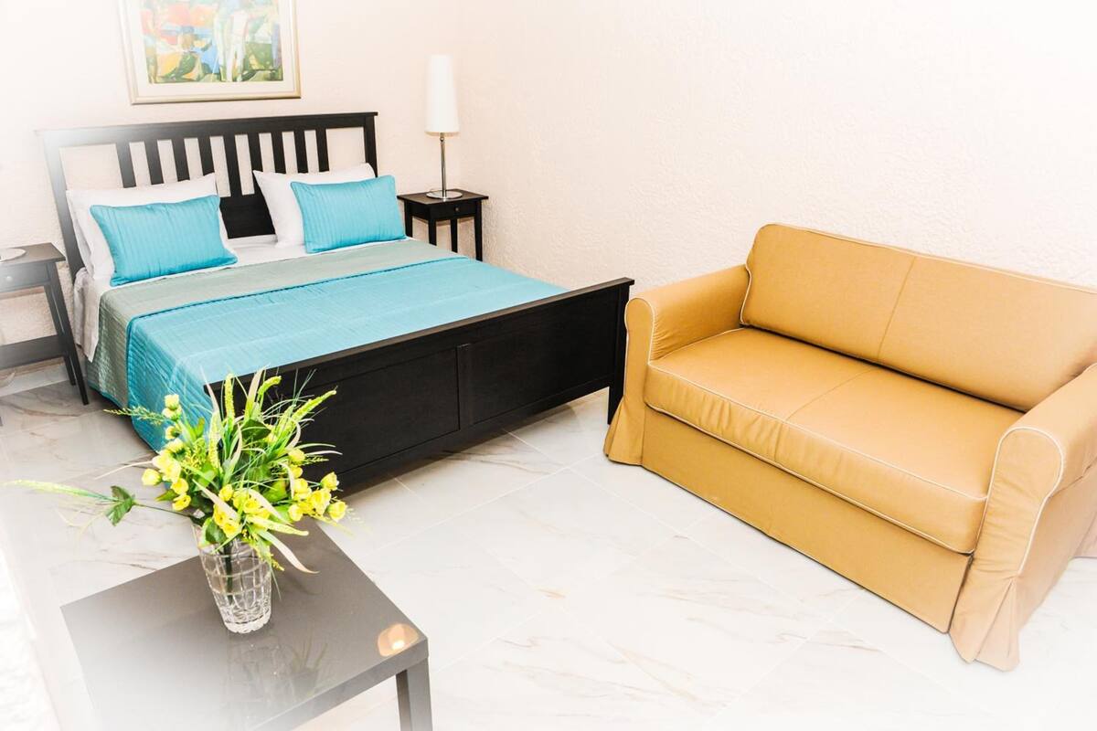 A bedroom with a bed, a nightstand, a lamp, a sofa, and a vase with flowers.