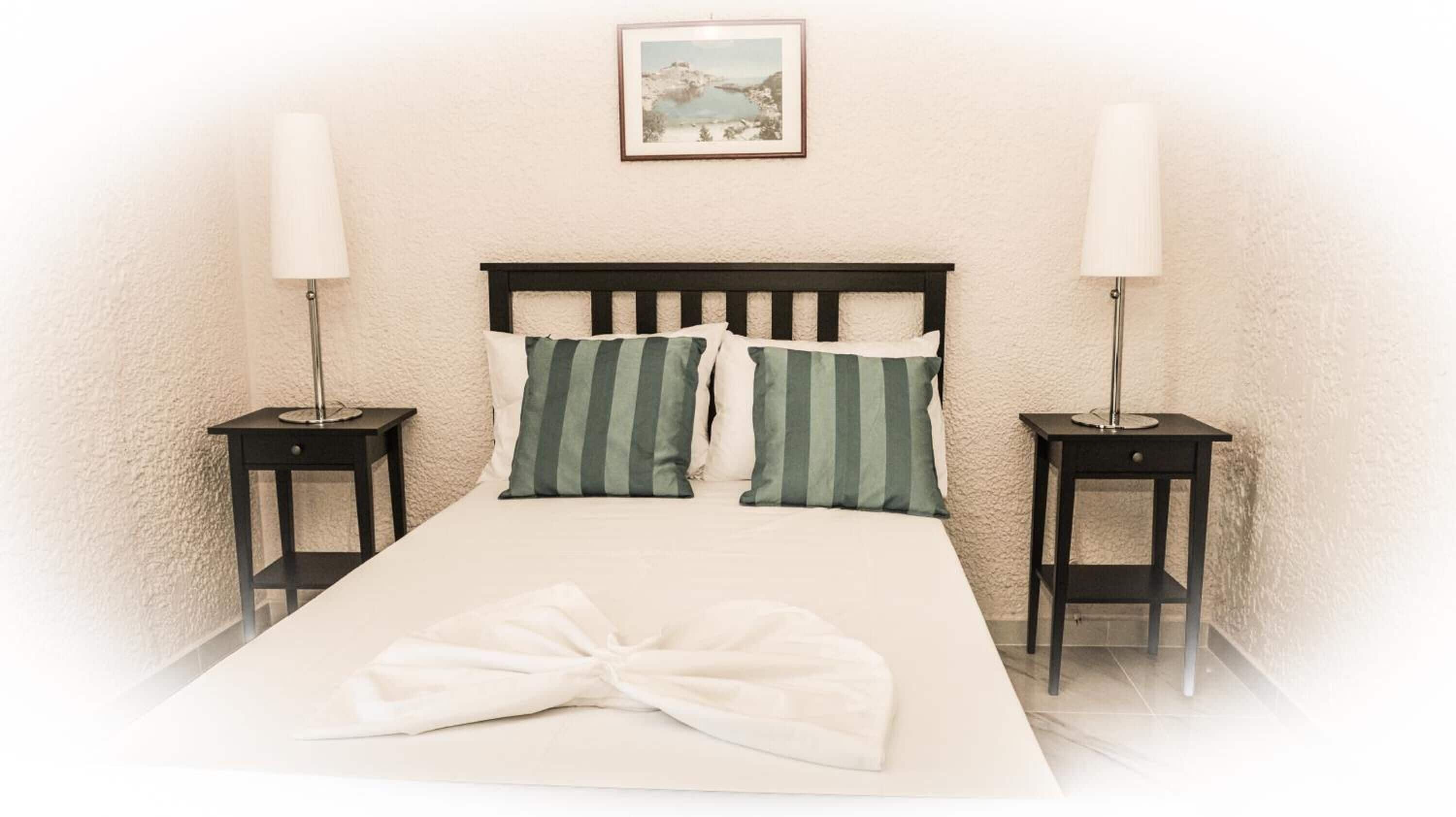 A neatly made bed with a white duvet and two pillows, flanked by two bedside tables with lamps, in a room with a textured wall and a framed picture above the bed.