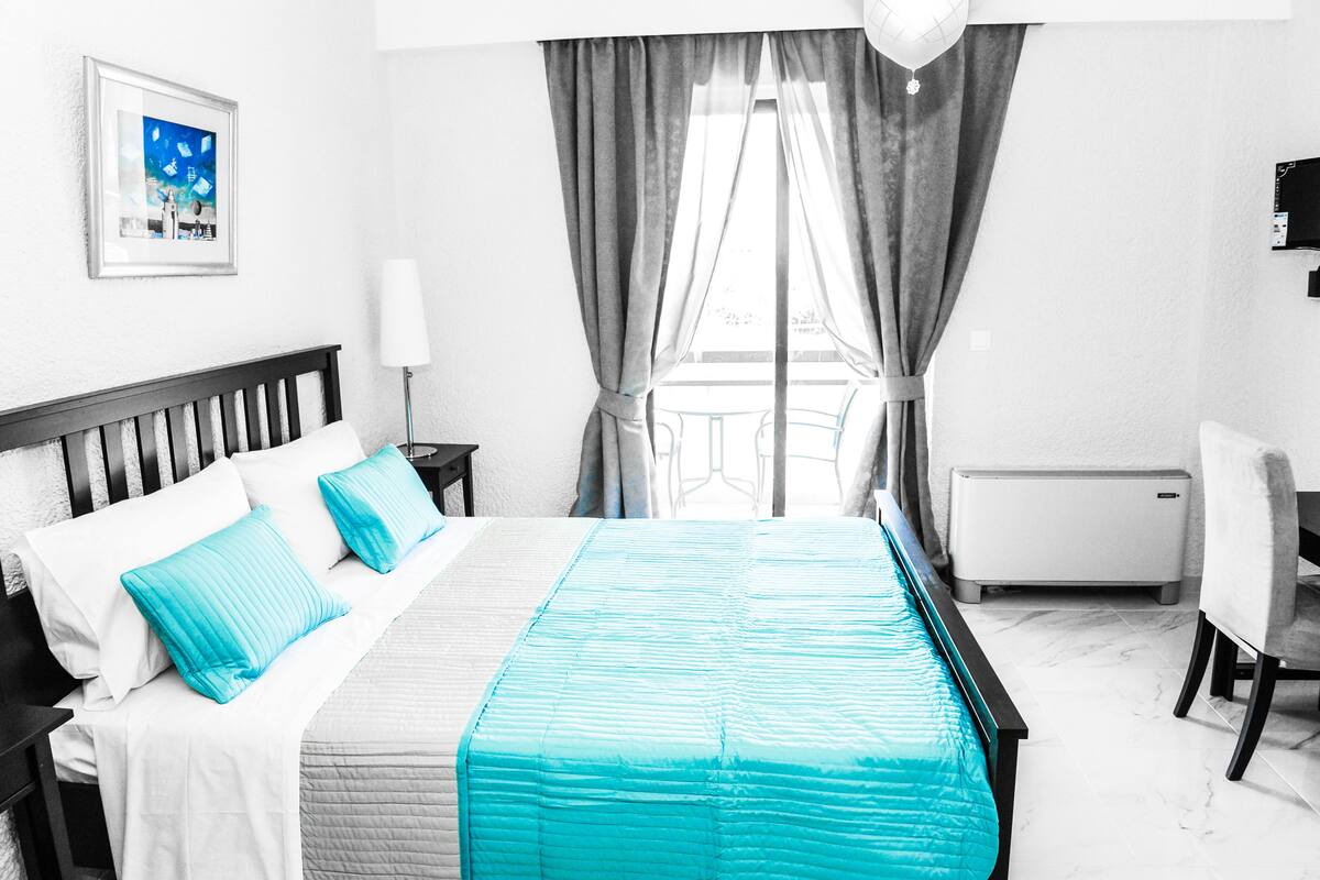 A hotel room with a bed, a desk, a chair, a window with curtains, a radiator, and a picture on the wall.