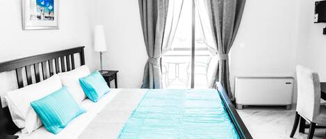 A hotel room with a bed, a desk, a chair, a window with curtains, a radiator, and a picture on the wall.