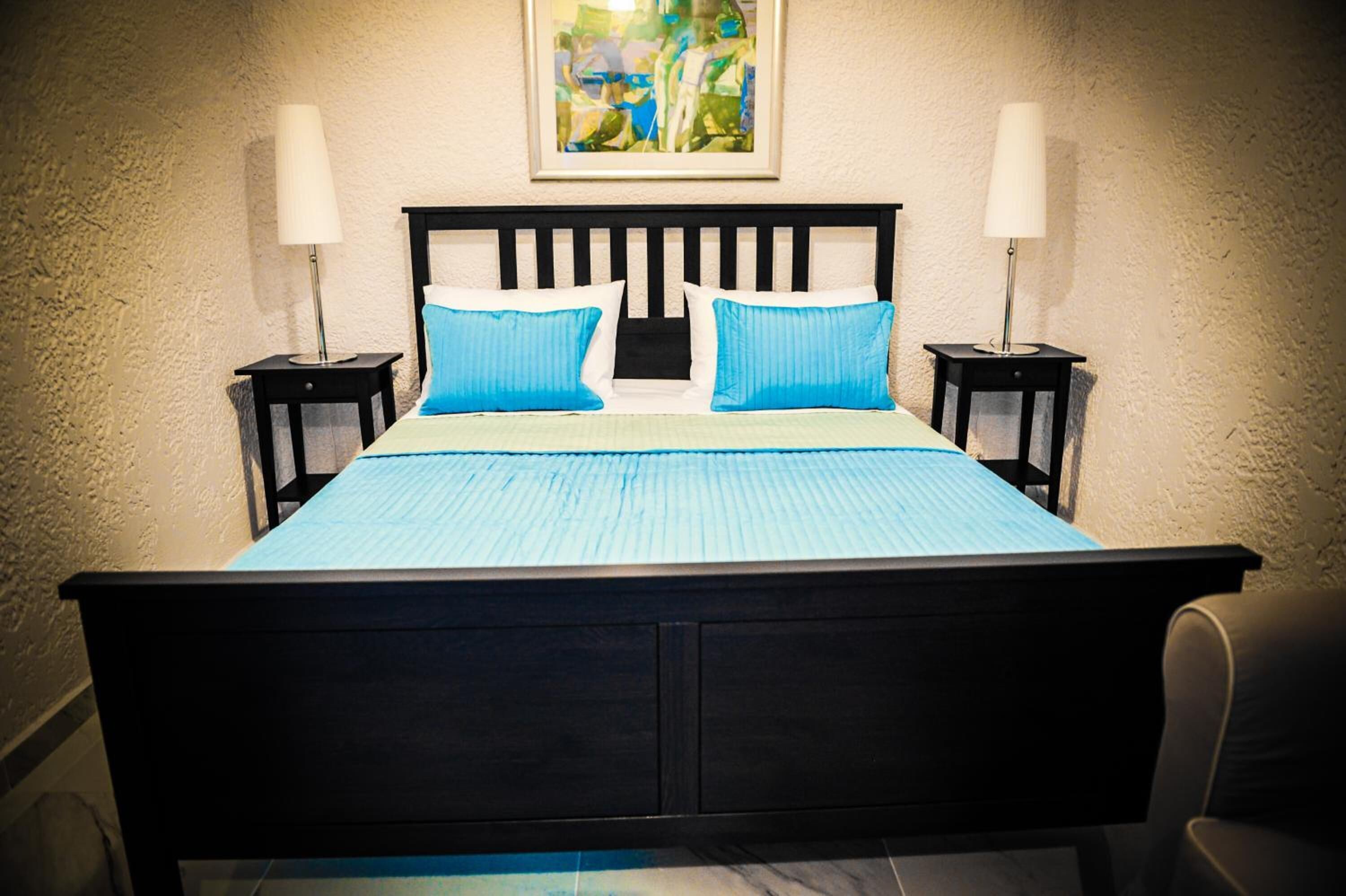 Ein Bett mit blauer Bettdecke und Kissen, seitlich von zwei Nachttischen mit Lampen begleitet, vor einer strukturierten Wand mit einem gerahmten Kunstwerk darüber.