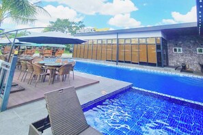 Une piscine avec des chaises longues et des tables, un bâtiment doté de grandes fenêtres et un panneau indiquant les horaires.