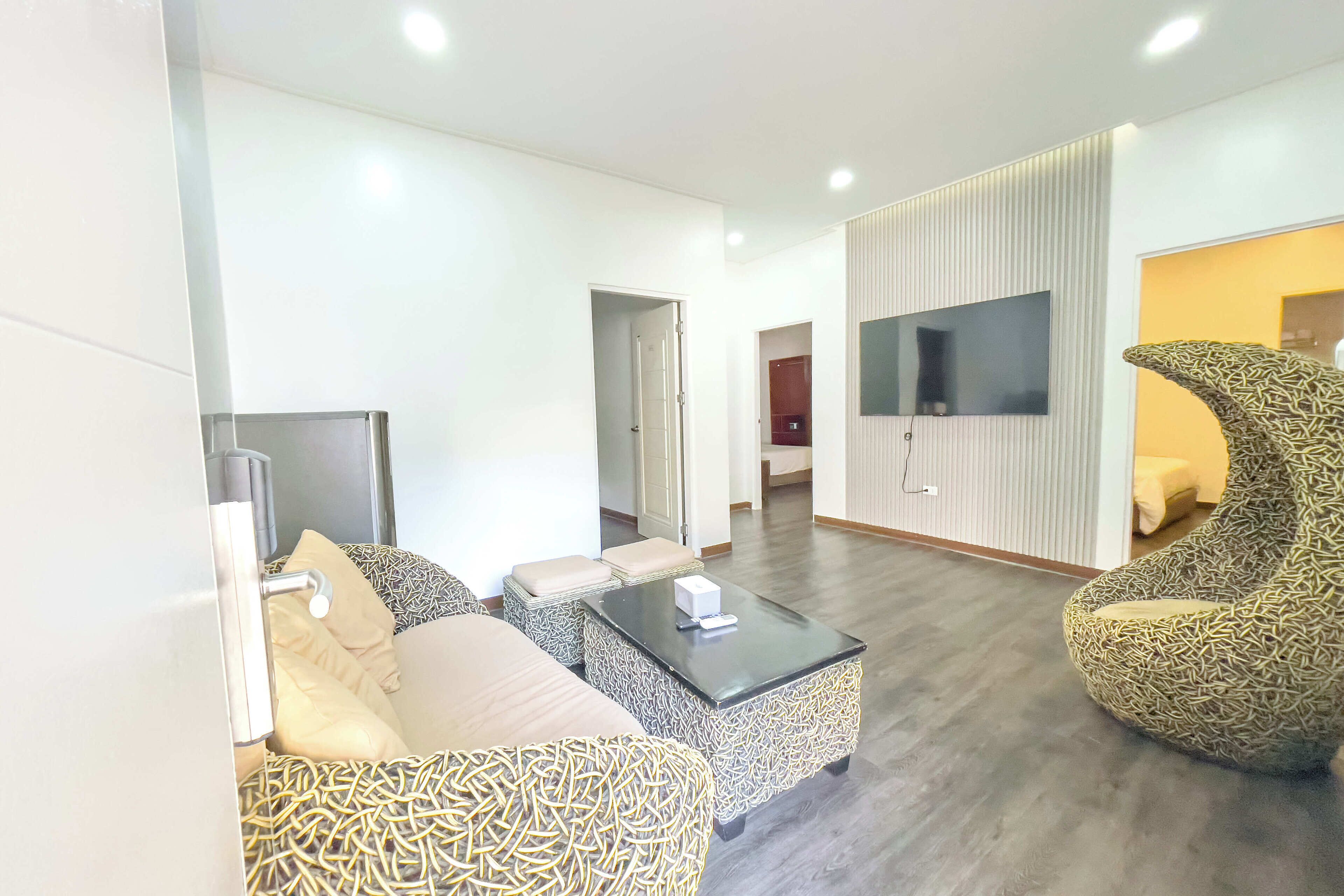 A modern living room with a sofa, ottoman, and a patterned armchair. A flat-screen TV is mounted on the wall, and there is a door leading to another room.