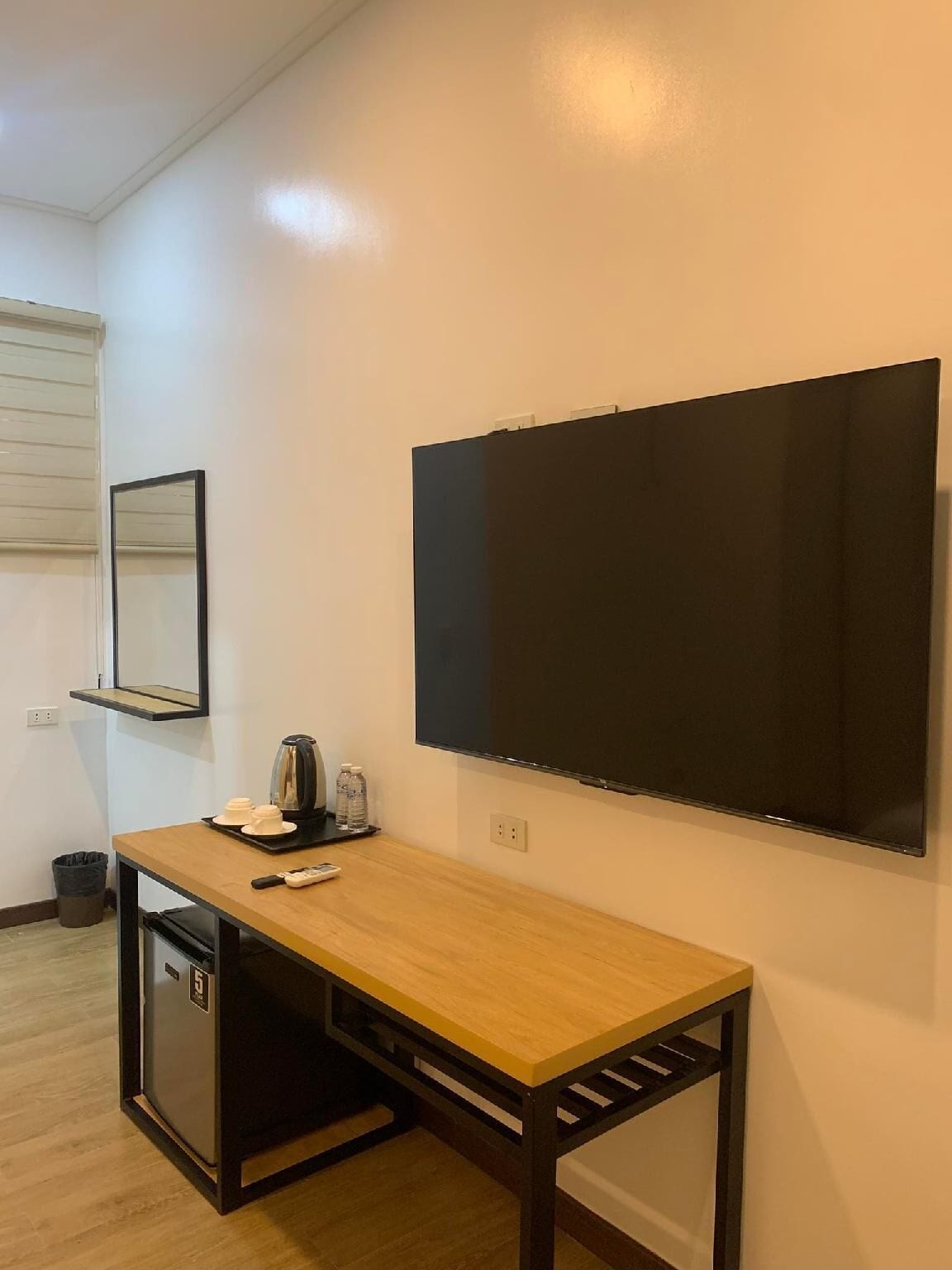 A compact room with a wooden desk, a small kitchenette, a flat-screen TV mounted on the wall, and a mirror above the desk.