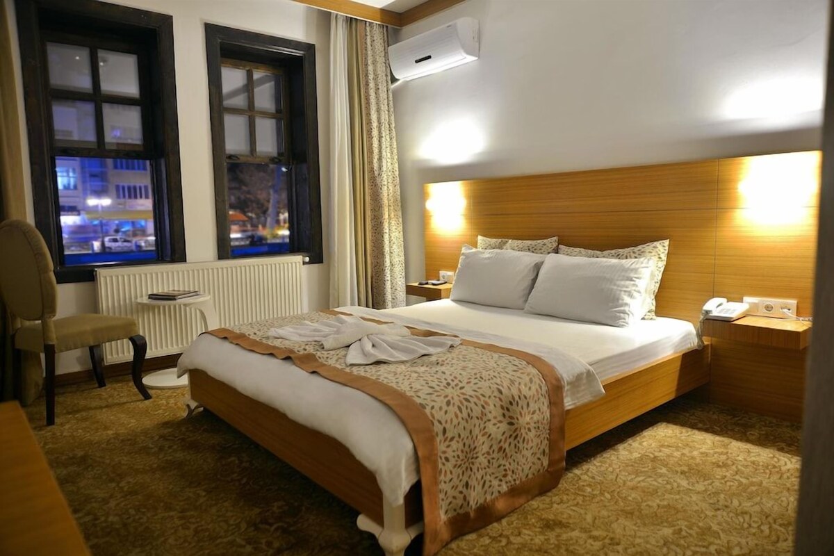 A hotel room with a large bed, a chair, a nightstand, a bedside table, and a window with a view of the city at night.
